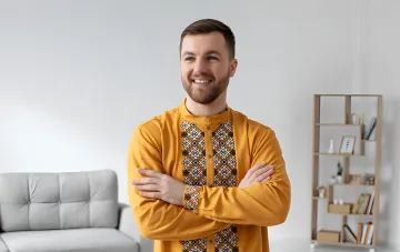 Усміхнений чоловік на ім'я Василь у жовтій сорочці з українським вишиванням, зі схрещеними руками, на тлі світлої вітальні — ілюстрація до статті про походження та характер імені Василь
