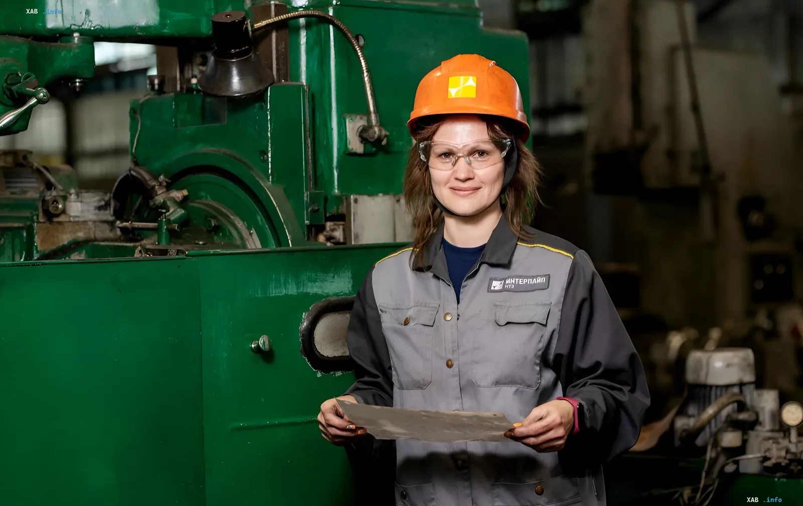 Frauen-Bedienungskraft bei Interpipe NTZ in Schutzhelm und Arbeitskleidung mit Metallteil an einer Maschine im Produktionsbereich