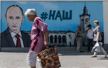 Bewohner von Berdjansk vor einem Graffiti mit Putins Porträt und #HAU-Hashtag vor zerstörten Gebäuden