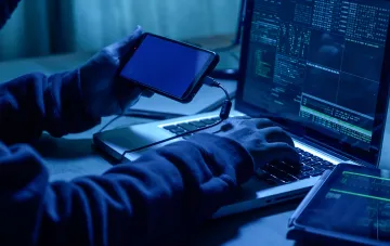 Хакер у капюшону підключає смартфон до ноутбука з потоком даних під час кібератаки на британські аеропорти, внаслідок якої були вкрадені дані 8,7 млн клієнтів