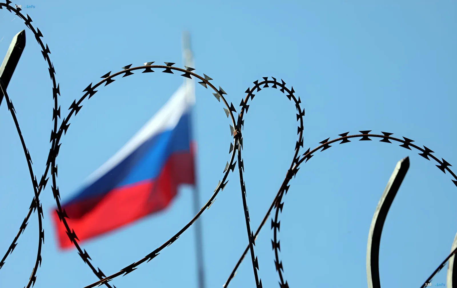 Колючая проволока на переднем плане и размытый российский флаг на фоне голубого неба — символ готовящегося осеннего пакета санкций ЕС против России