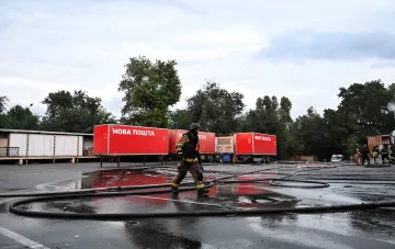 Feuerwehrmann unter ausgelegten Schläuchen am zerstörten Fulfillment-Zentrum von Nova Poshta in Swjatopetriwka, im Hintergrund rote Container des Unternehmens