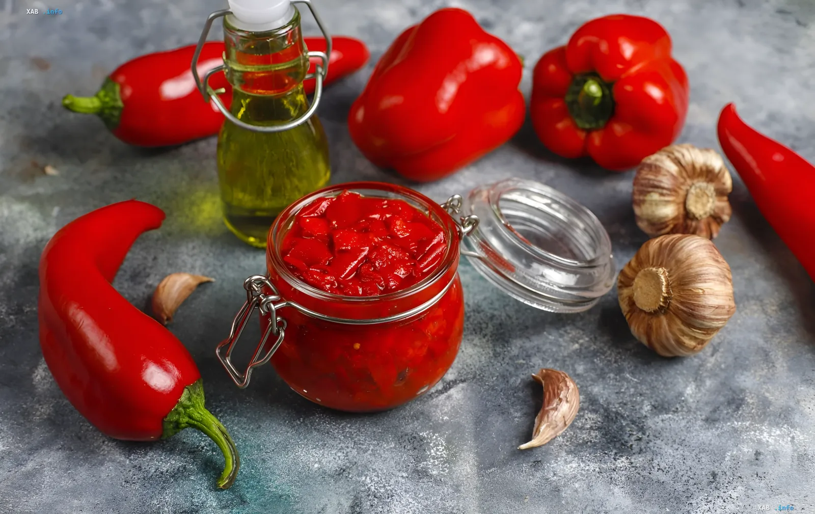 Стеклянная банка с маринованным красным перцем на зиму с чесноком, свежие болгарские и острые перцы, головки чеснока и масло на сером каменном фоне — мясистая заготовка по правилу 6 минут
