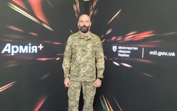 Артем Романюков в военной форме на фоне логотипов Министерства обороны Украины и Армія+