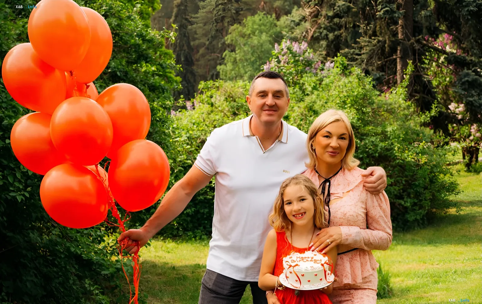 Семья в парке: мужчина с оранжевыми шарами, женщина и девочка с праздничным тортом — подготовка к первому звонку