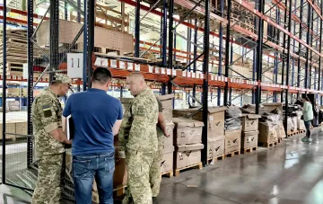 Cargo inspection at a warehouse terminal: military inspectors and a staff member with documents examine pallets of boxes — an example of customs delays and financial risk for business