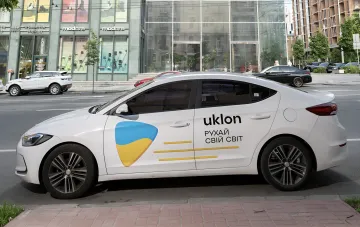 White Uklon taxi car with the company logo and the Ukrainian flag on the door, parked on a street in Kyiv — the company explains its stance against a taxi price cap amid the alert regime debate