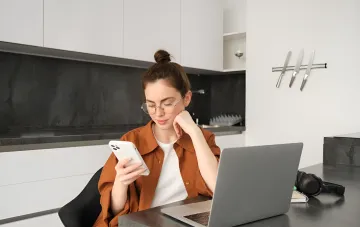 Женщина в очках сидит за ноутбуком и смотрит в белый смартфон, иллюстрирующая тему общения в рабочих чатах ОСМД и требования языкового омбудсмена к использованию украинского языка
