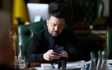 Владимир Зеленский за рабочим столом в кабинете, изучающий смартфон на фоне украинского флага — в контексте внесения в Раду законопроекта об уголовной ответственности за дроп и мошенничество с платежными инструментами