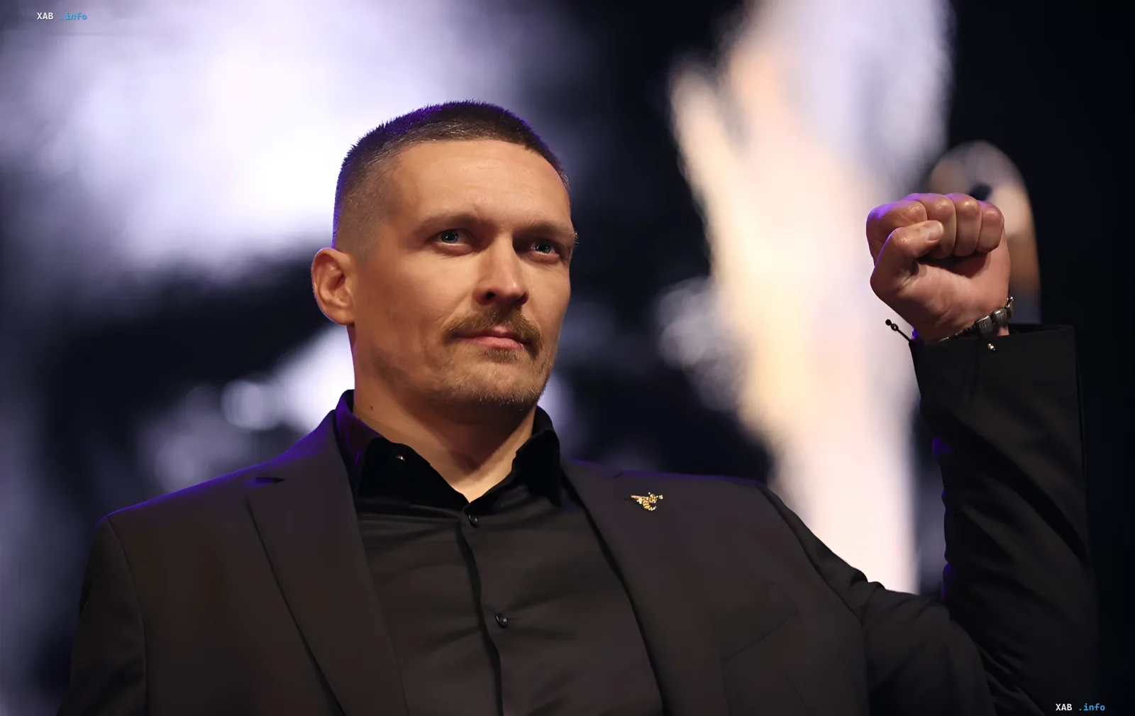 Боксер Александр Усик в черном костюме с поднятой рукой на фоне арены перед боем с Уайлдером в Лас-Вегасе
