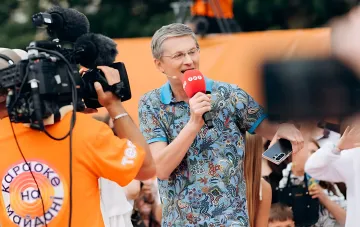 Ведучий шоу «Караоке на майдані» з мікрофоном під час зйомок, оператор з камерою на тлі помаранчевого банера проєкту