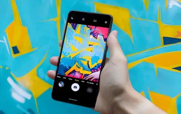 Ручка держит Google Pixel 11 с открытой камерой, демонстрирующей активный режим AI и настройки Android 17 на фоне яркой уличной стены
