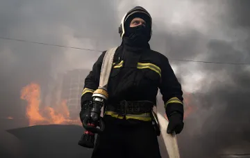 Пожарный в защитной экипировке со стволом рукава на фоне дыма и пламени после ночного удара по промышленному предприятию в Каменском