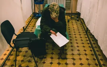 Военнослужащий в камуфляжной форме и кепке сидит в медицинском кабинете и изучает документ, связанный с присвоением статуса участника боевых действий