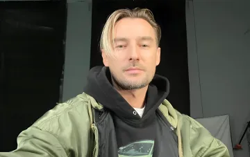 Редкое домашнее фото из публикации Алана Бадоева: мужчина с платиновыми волосами в оливковой куртке и чёрном худи на фоне тёмного интерьера