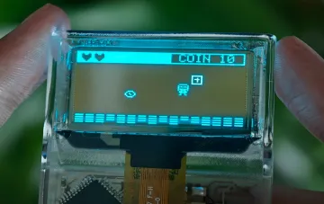 Transparente Retro-Konsole Arduview im Hosentaschenformat mit sichtbarer Platine und OLED-Display, auf dem ein Pixel-Art-Spiel läuft