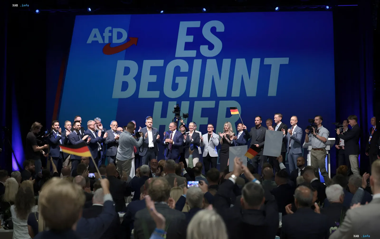 Съезд партии AfD в Германии: политики на сцене с немецкими флагами перед аудиторией, лозунг «ES BEGINNT HIER»