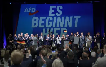 Съезд партии AfD в Германии: политики на сцене с немецкими флагами перед аудиторией, лозунг «ES BEGINNT HIER»