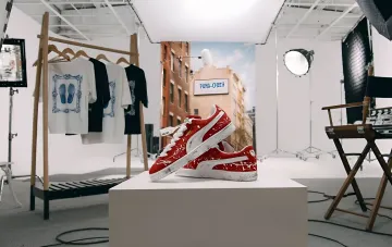 Красные кроссовки PUMA Suede с эффектом потертости и брызгами краски на подиуме в фотостудии, на фоне баннер PUMA x CHER'17 и вешалка с футболками коллекции