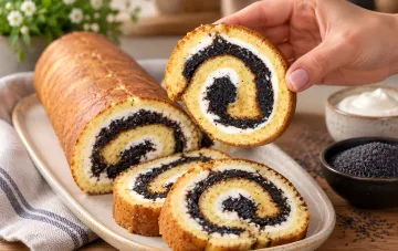 Geschnittener Sechs-Löffel-Rollenkuchen mit Mohn- und Quarkfüllung, ein Stück wird von einer Hand gehalten, daneben Schalen mit Mohn und Creme