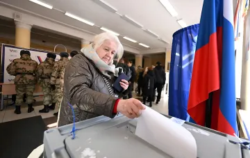 Женщина опускает бюллетень в урну на псевдовыборах на оккупированной территории, рядом российский флаг и военные