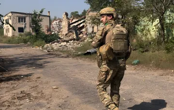 Білець Сил оборони України в камуфляжі та тактичному спорядженні на тлі зруйнованої будівлі