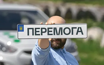 Мужчина держит украинский автомобильный номер «ПЕРЕМОГА» на фоне машины — напоминание МВД о необходимости пластикового водительского удостоверения вместо «Діі»