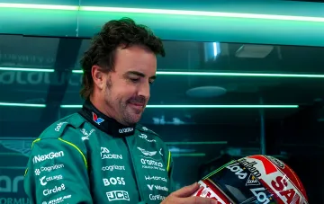 Фернандо Алонсо в форме команды Aston Aramco Formula One Team держит свой гоночный шлем на фоне неоновых огней боксов