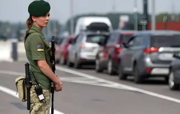 Armed Ukrainian Border Guard officer with rifle and baton stands at a checkpoint, cars in a queue in the background