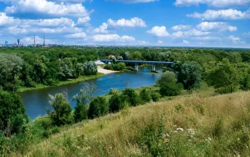 Landschaft der Ostukraine: ein Fluss, eine Brücke und grüne Hügel vor industriellen Anlagen – ein Gebiet, dessen Erinnerung das digitale Projekt MemoriDim bewahrt