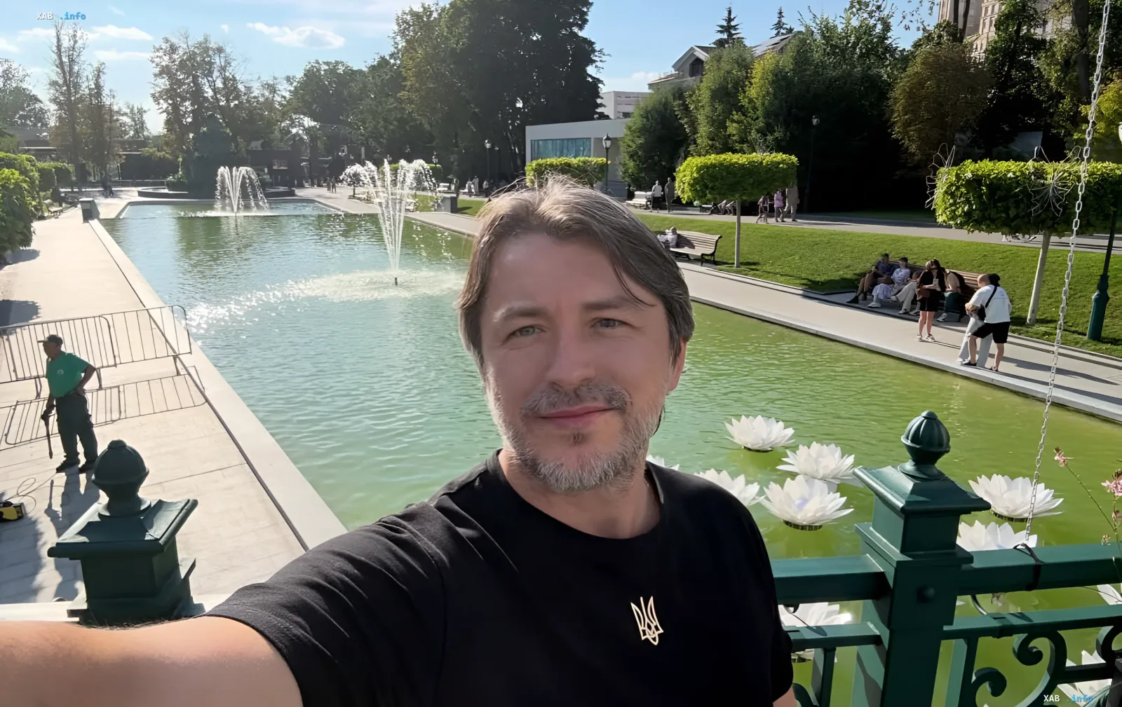 Сергій Притула на особистому селфі на тлі паркового ставка з фонтанами та білими лотосами в чорній футболці з золотим символом