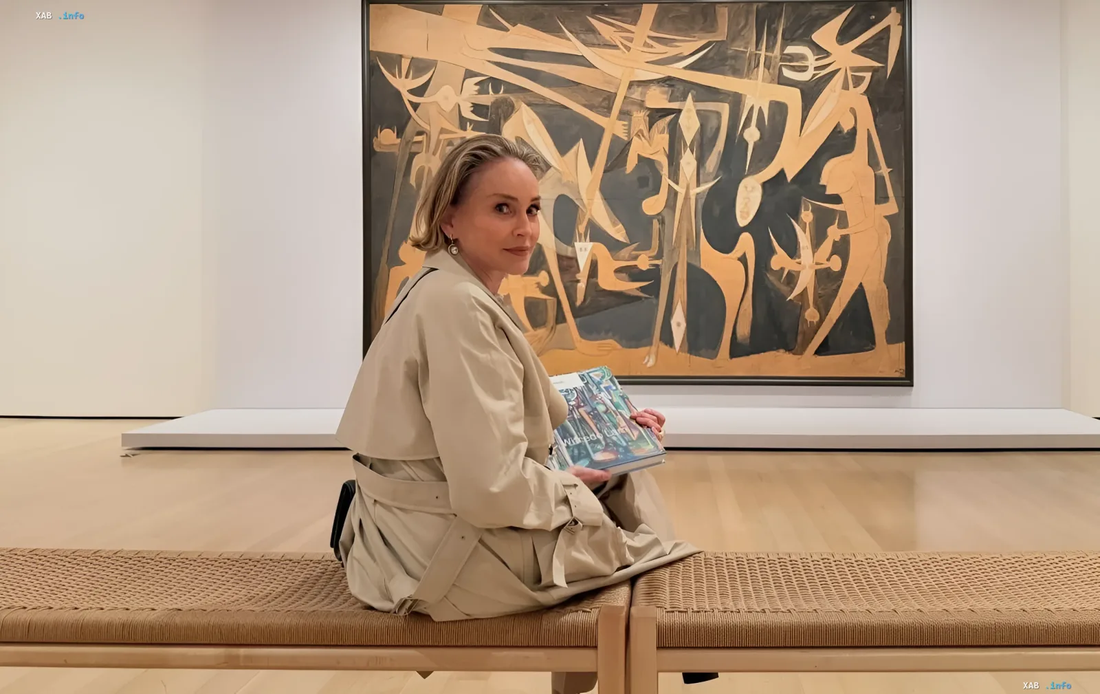 Шэрон Стоун у бежевому тренчкоті сидить на лавці в галереї з книгою «Поштовий індекс Україна» на тлі абстрактного полотна