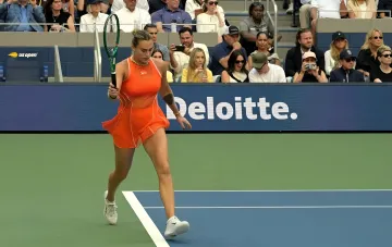 Арина Соболенко в оранжевом платье на корте US Open во время финального матча, на фоне трибун и рекламы Deloitte