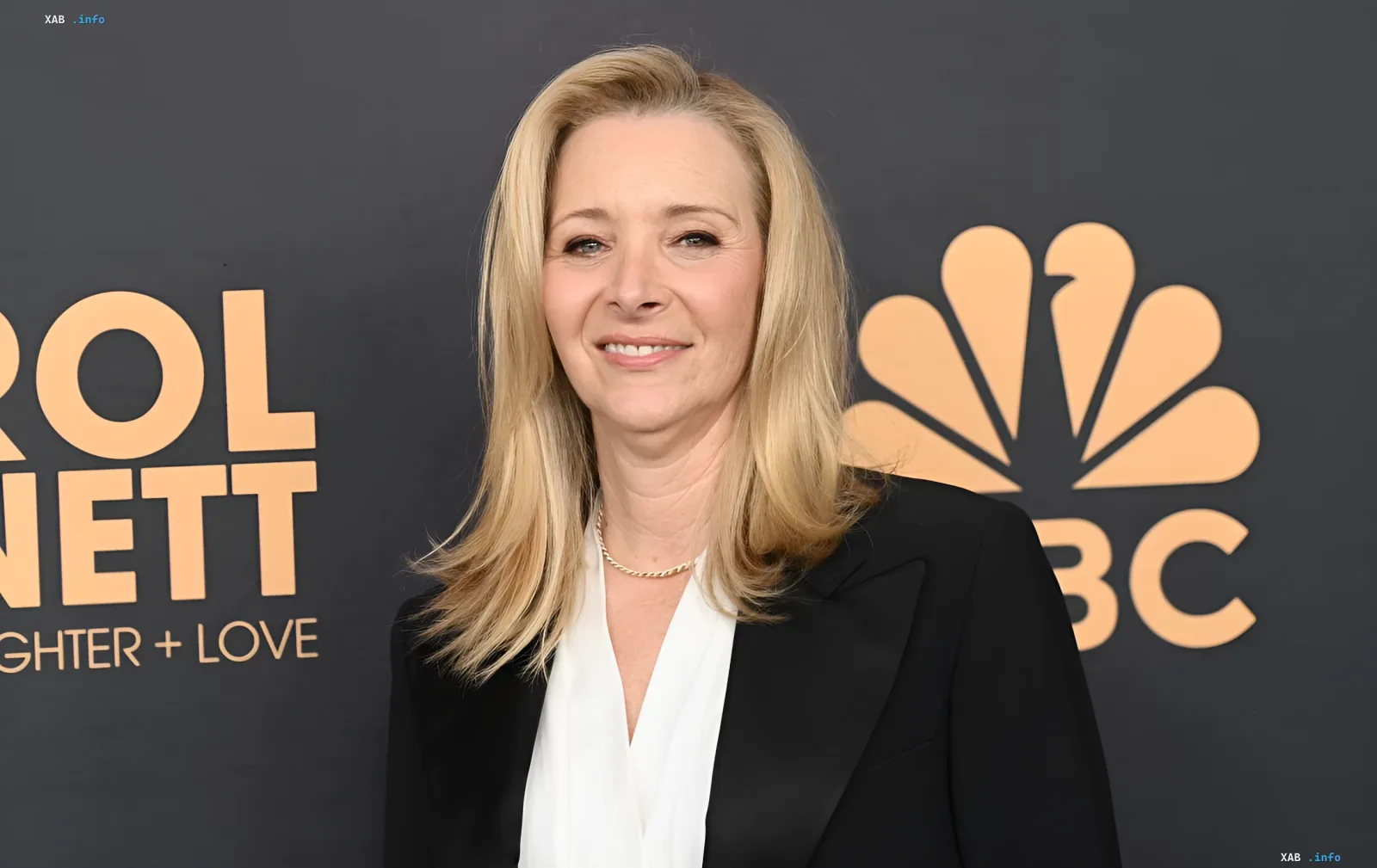Лиза Кудроу в чёрном пиджаке и белой блузке на фоне логотипа NBC — образ, который она выбрала для выхода на «Эмми» вместе с сыном