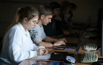 Oberstufenschüler in einem Computerraum: Ein Mädchen macht Notizen im Heft, ein Junge arbeitet am Laptop – Illustration zur neuen profilorientierten Lehrform in ukrainischen Lyzeen