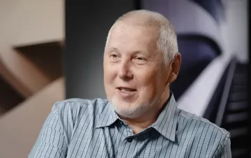 Historian Vitaliy Dribnytsia in a checked shirt against a modern interior backdrop; he has taken a break from on-air work due to cancer treatment in a Berlin suburb
