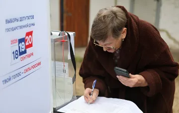 Пожилая избирательница заполняет документ на участке по выборам в Госдуму рядом с прозрачным ящиком для голосования и плакатом с датами 18–20 сентября