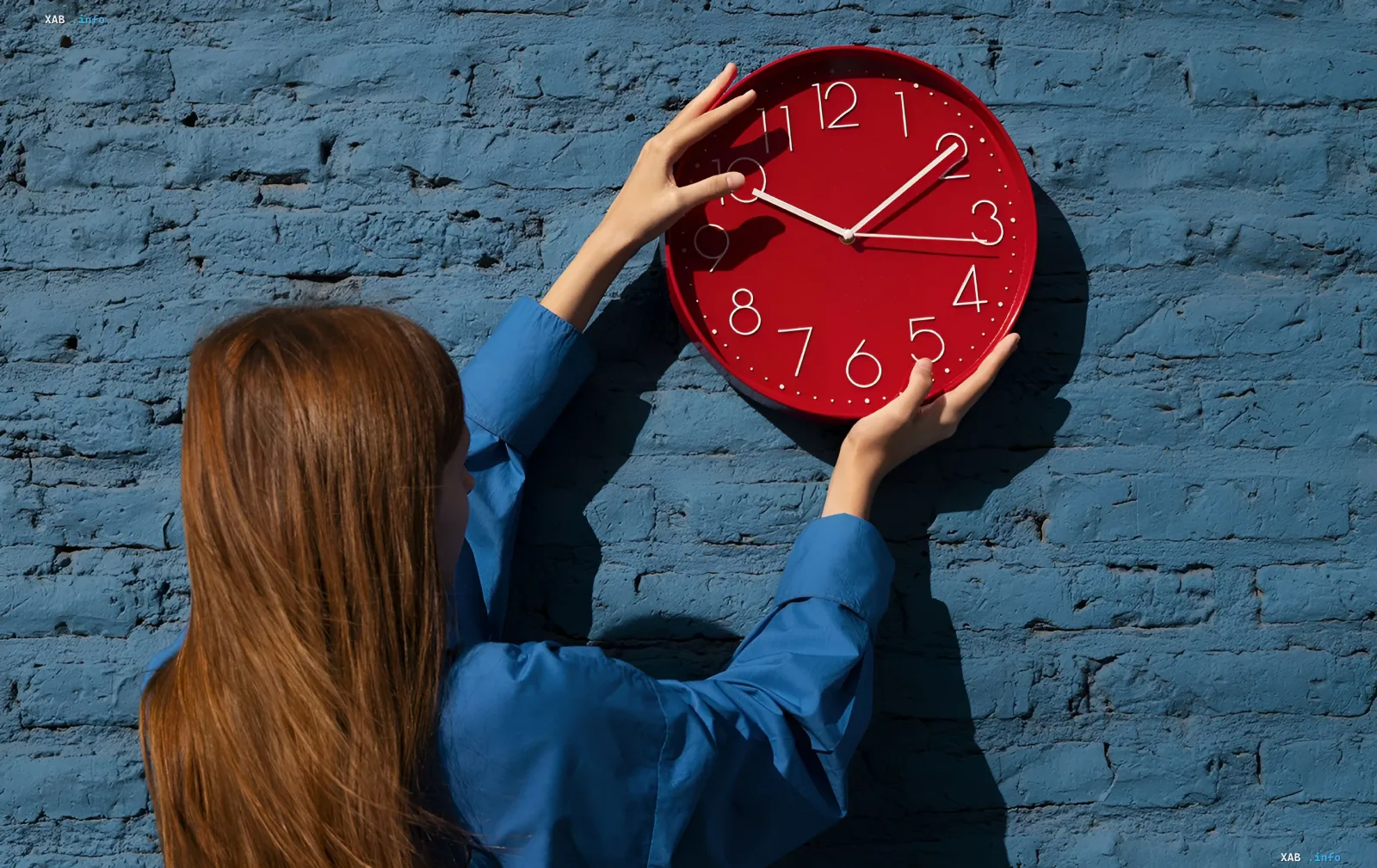 Woman holding a red wall clock against a blue brick wall — symbol of switching to winter time in Ukraine and the EU on 25 October 2026