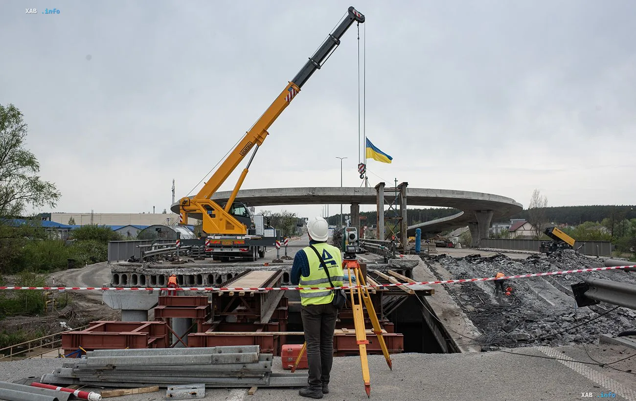 Baustelle zum Wiederaufbau einer zerstörten Überführung in der Ukraine: Ein Kran hebt die ukrainische Flagge, während ein Ingenieur Vermessungsarbeiten durchführt — Symbol des großangelegten Infrastrukturaufbaus im Rahmen der Vergabereform