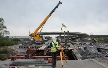 Reconstruction site of a destroyed overpass in Ukraine: a crane lifts the Ukrainian flag while an engineer takes survey measurements — a symbol of large-scale infrastructure rebuilding under the procurement reform