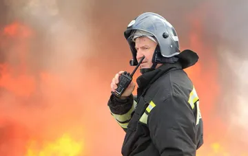 Firefighter in a protective helmet coordinates via radio against a backdrop of flames after a strike on a Russian oil refinery