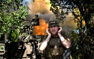 Ukrainian soldier in camouflage covering ears during fire from an anti-aircraft gun against an explosion background
