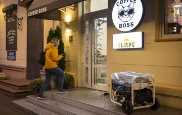 Frau in gelber Jacke vor einem Café mit laufendem Generator, der Strom nach einem Blackout liefert