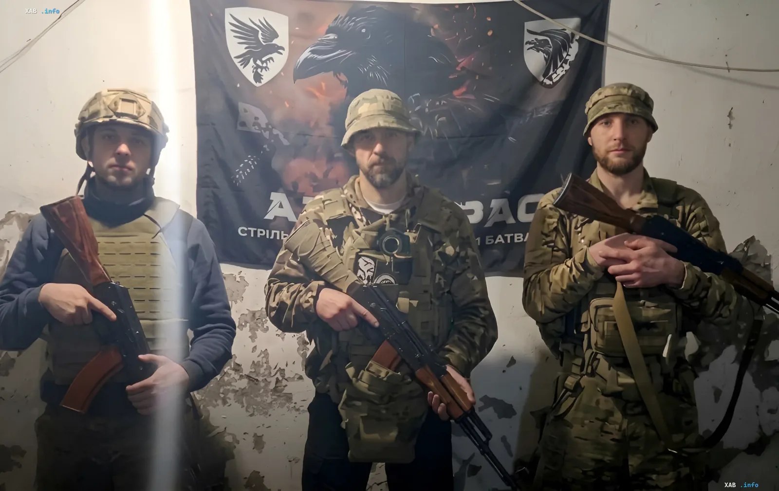 Трое украинских военных в камуфляже и с оружием на фоне флага с изображением ворона