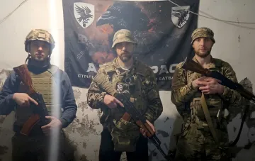 Трое украинских военных в камуфляже и с оружием на фоне флага с изображением ворона