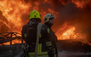 Feuerwehrleute in Schutzhelmen blicken auf ein Großfeuer nach einem Drohnenangriff