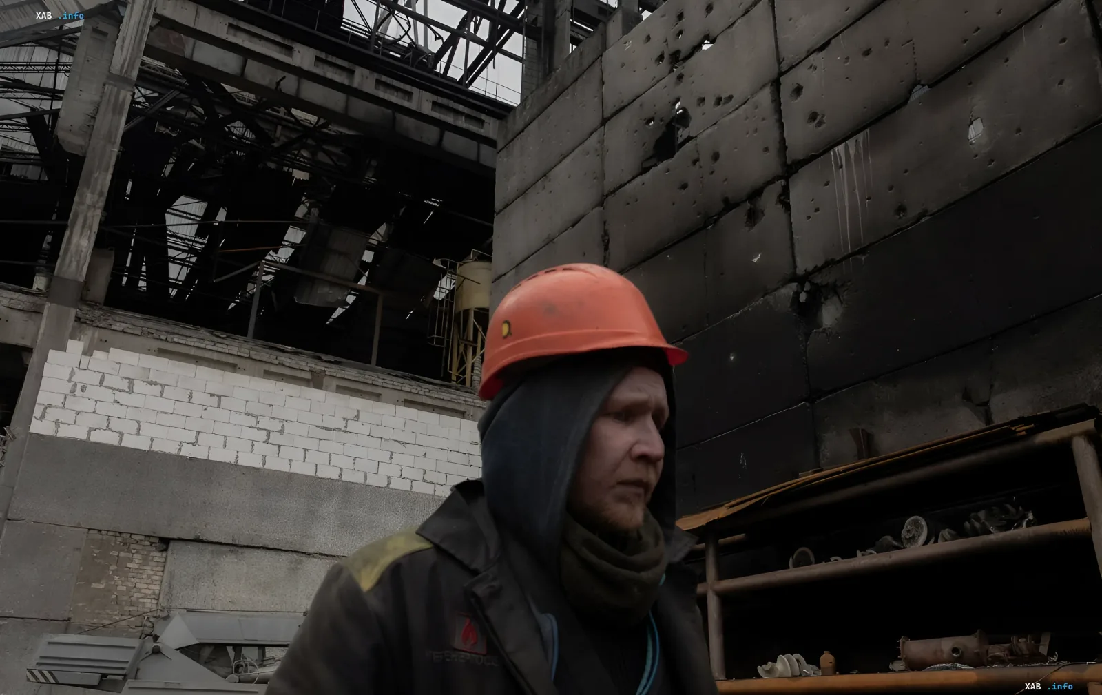 A worker in an orange hard hat against the backdrop of a destroyed and damaged industrial facility with bullet and shrapnel marks
