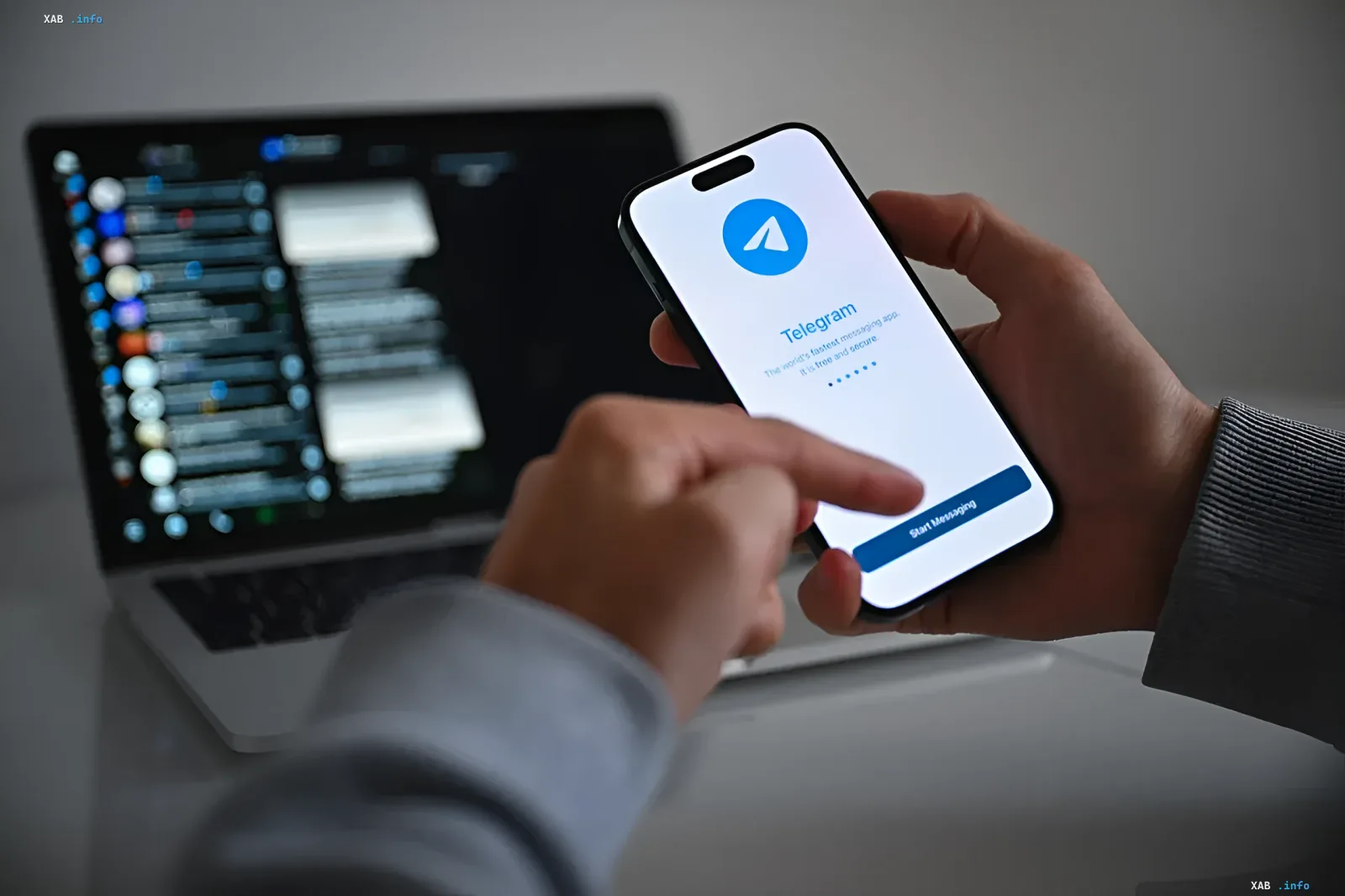 Человек держит в руках смартфон с логотипом Telegram на экране рядом с открытым ноутбуком