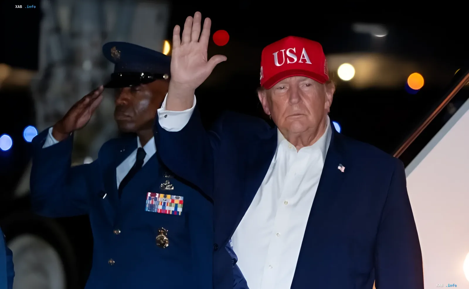 Дональд Трамп в красной кепке USA машет рукой рядом с военным в форме
