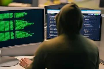 Хакер в капюшоне сидит за компьютером с исходным кодом в темноте